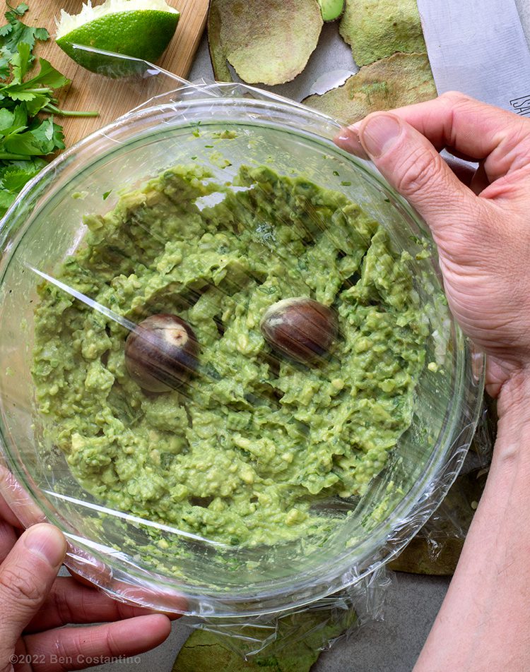 guacamole in a bowl being stored with two avocado pits and covered with saran wrap
