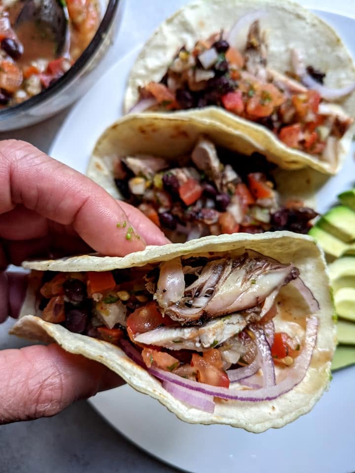 a plate of jerk chicken tacos, holding up a taco with chopped jerk chicken, onions, and salsa with a side of sliced avocado